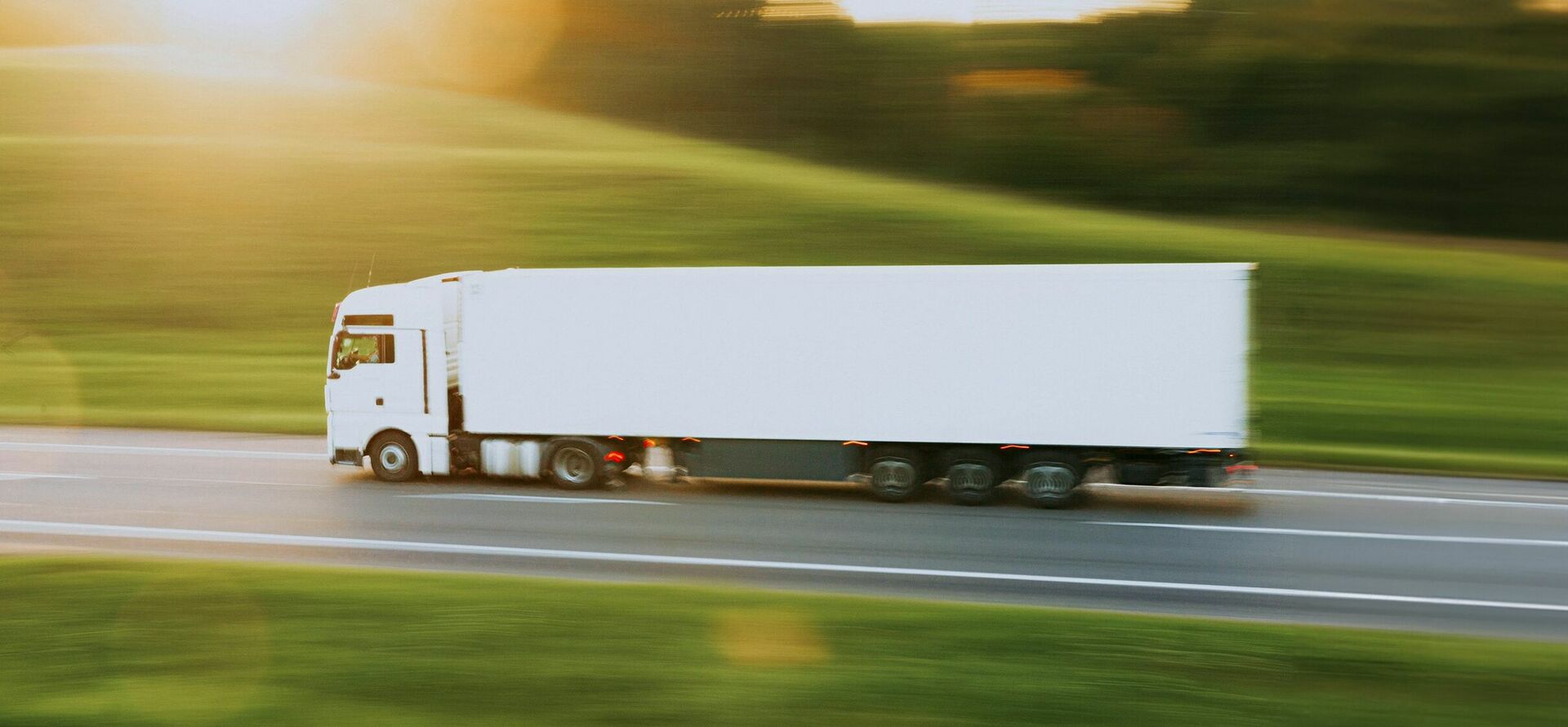 Weißer LKW, der auf einer Straße fährt, seitlich gesehen vor einer untergehenden Sonne und grüner Landschaft.